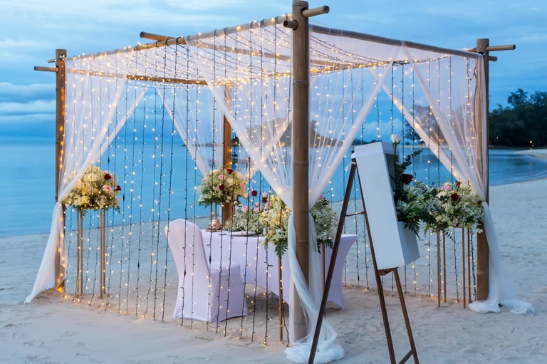 Romantic beach marriage proposal setup with wooden structure adorned with fairy lights and floral decorations, set against ocean backdrop during sunset