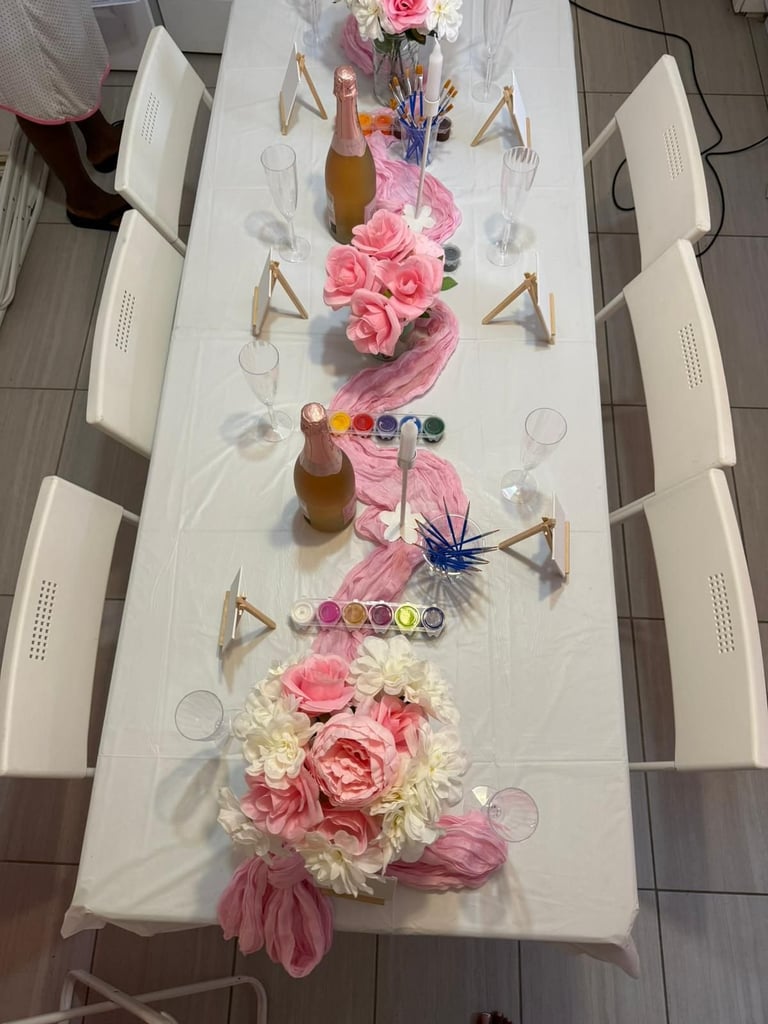 Overhead view of a white dining table with pink flower arrangements, champagne bottles, and art supplies arranged down the center