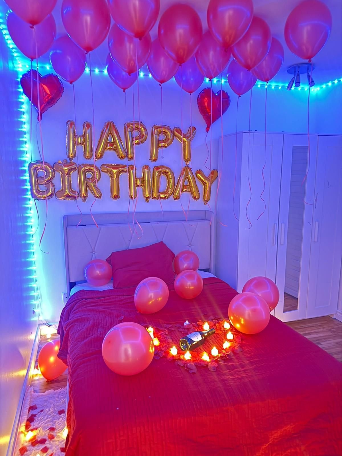 Bedroom decorated for birthday with pink and purple balloons, golden Happy Birthday sign, neon lights, and glowing string lights on bed