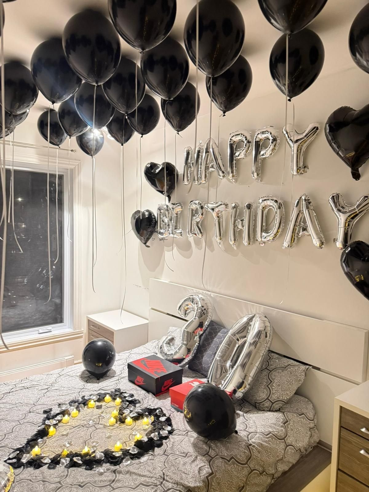 A bedroom decorated for a birthday with black and silver balloons, HAPPY BIRTHDAY letter balloons, rose petals on the bed, and gift boxes