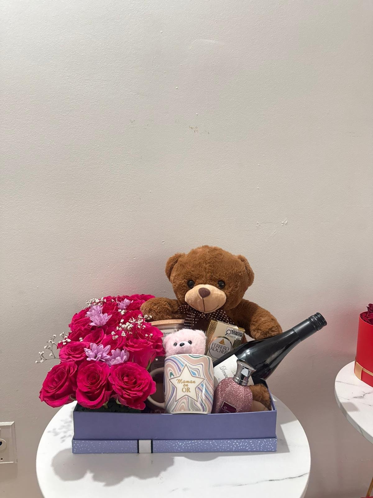 Gift box arrangement with pink roses, teddy bear, candle, wine bottle on white table